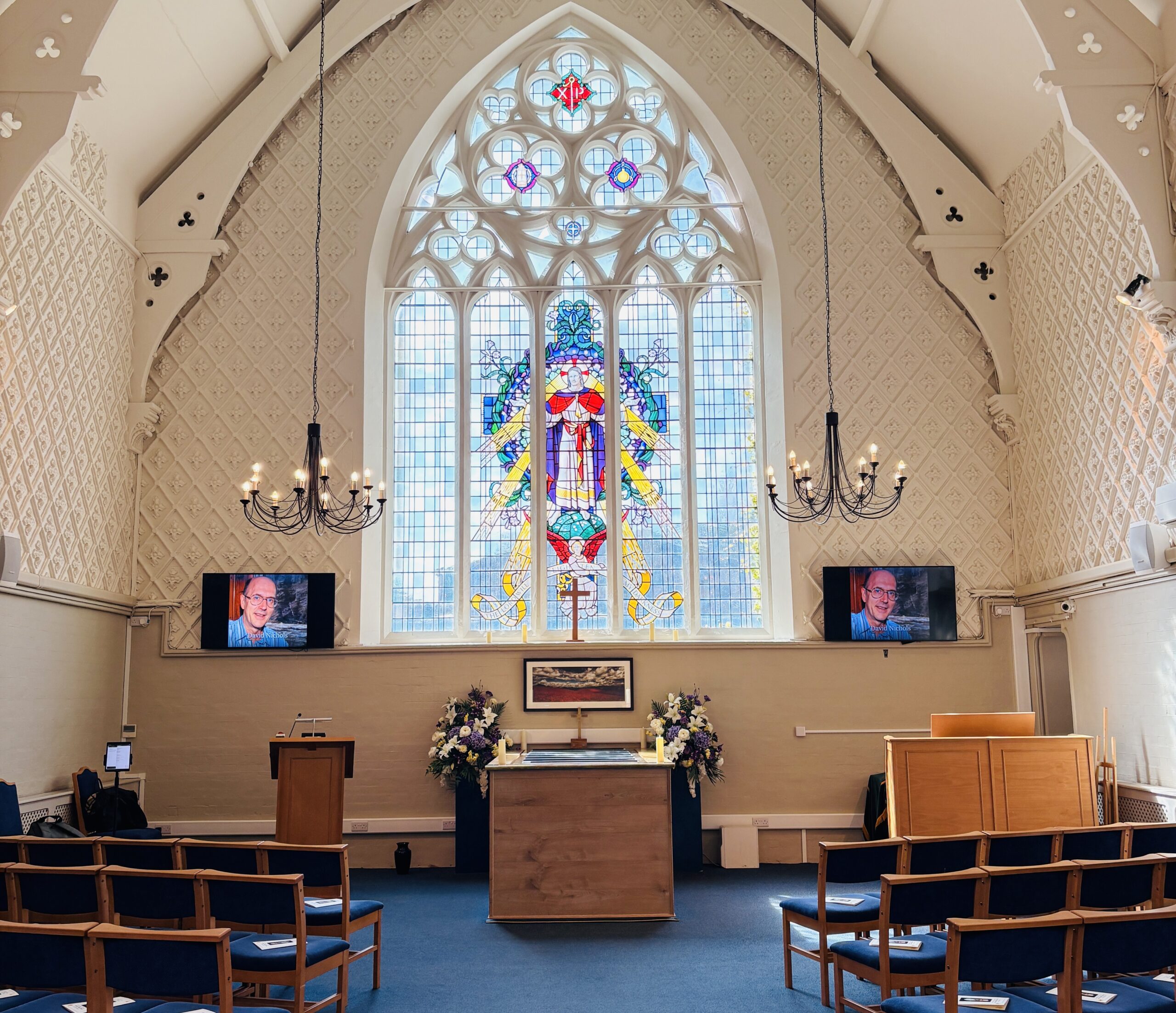 Photo of the chapel at Beckenham Crematorium, featuring a beautiful stained glass window, high ceilings with beautiful chandelier lights dangling from the ceiling.
